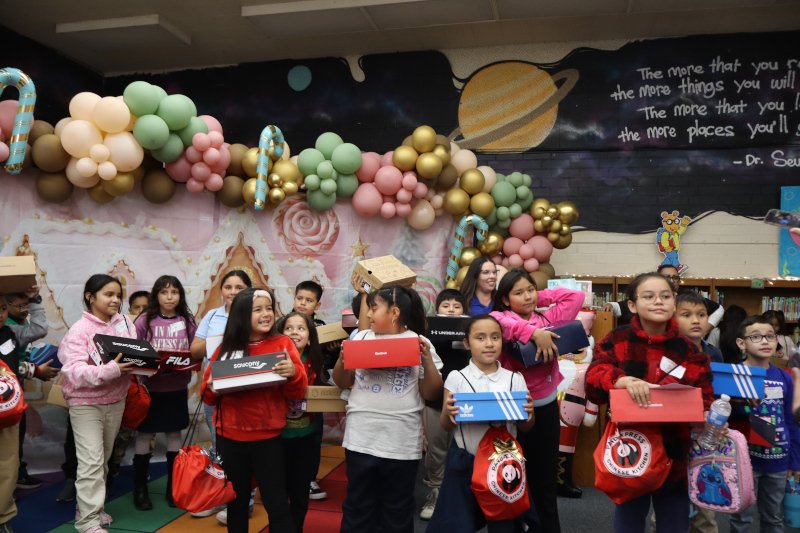 Elementary school students stand together indoors holding Panda Express swag bags and shoe boxes during a holiday giveaway