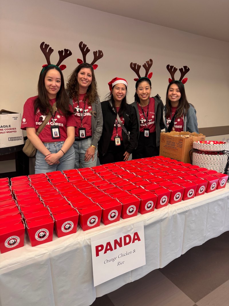 Panda Cares volunteers wearing festive reindeer headbands stand behind a table filled with neatly arranged red Panda Express meal boxes at a holiday community event
