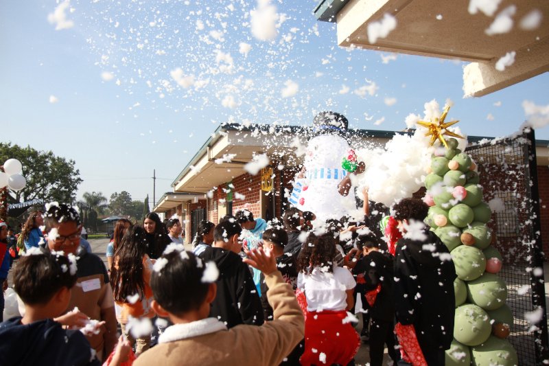 Children and adults gathered outdoors as white foam fills the air during a festive celebration near decorated buildings and balloon displays